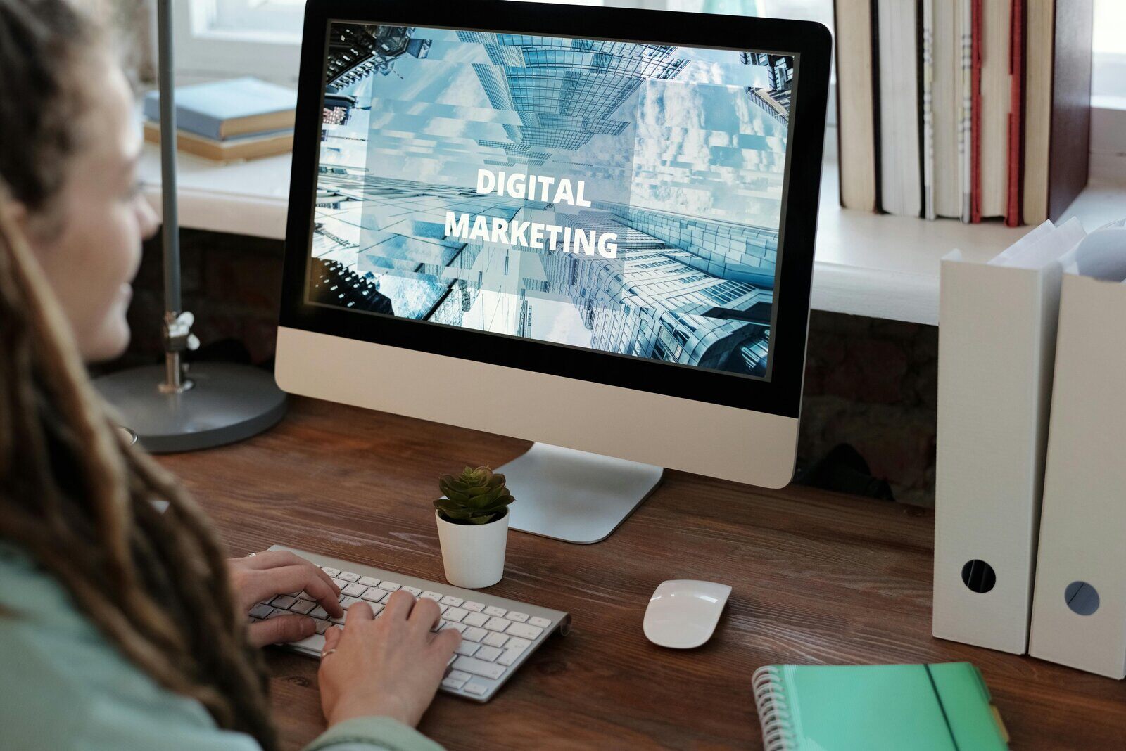 woman typing on a keyboard at a wooden desk with -Digital Marketing- displayed on an iMac screen, emphasizing the importance of online strategies for builders. Builder Funnel